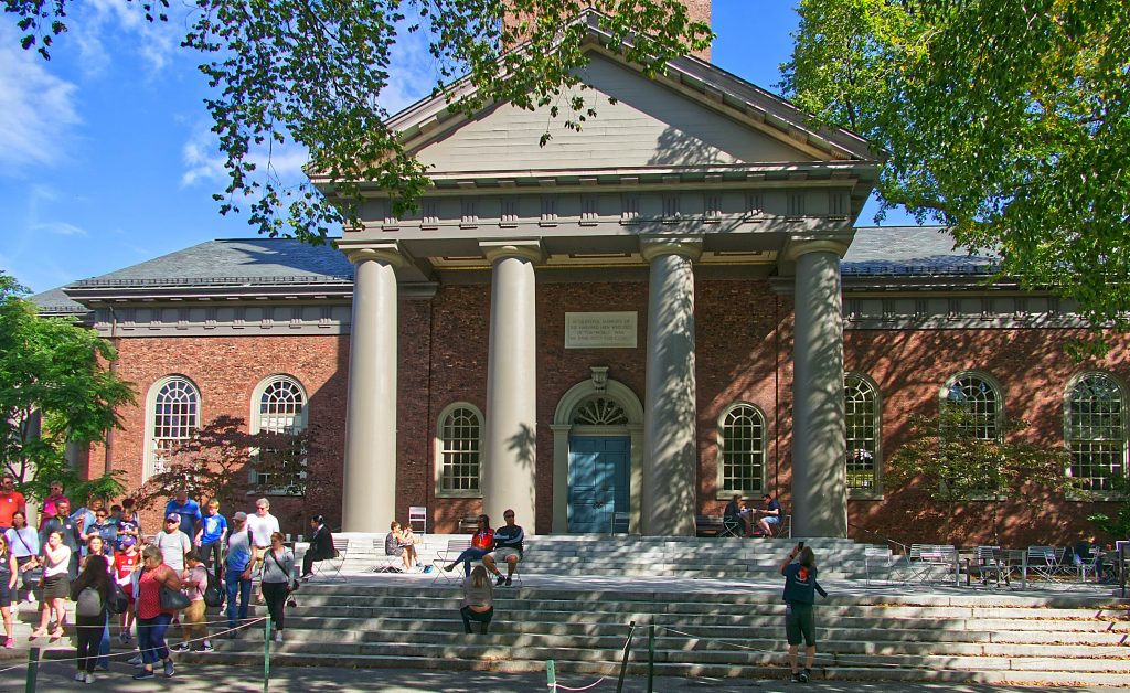 A crowd of people outside a majestic brick building, possibly Harvard, discussing the admission criteria for hopeful applicants.