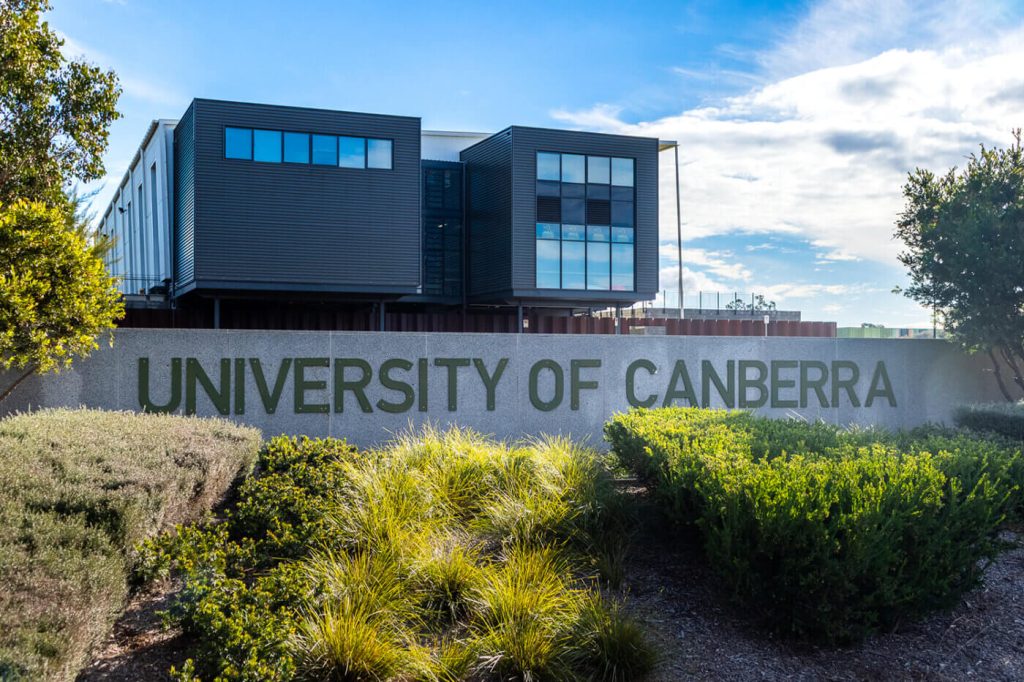 University of Canberra sign with blue lettering and a kangaroo emblem in front of a modern building.